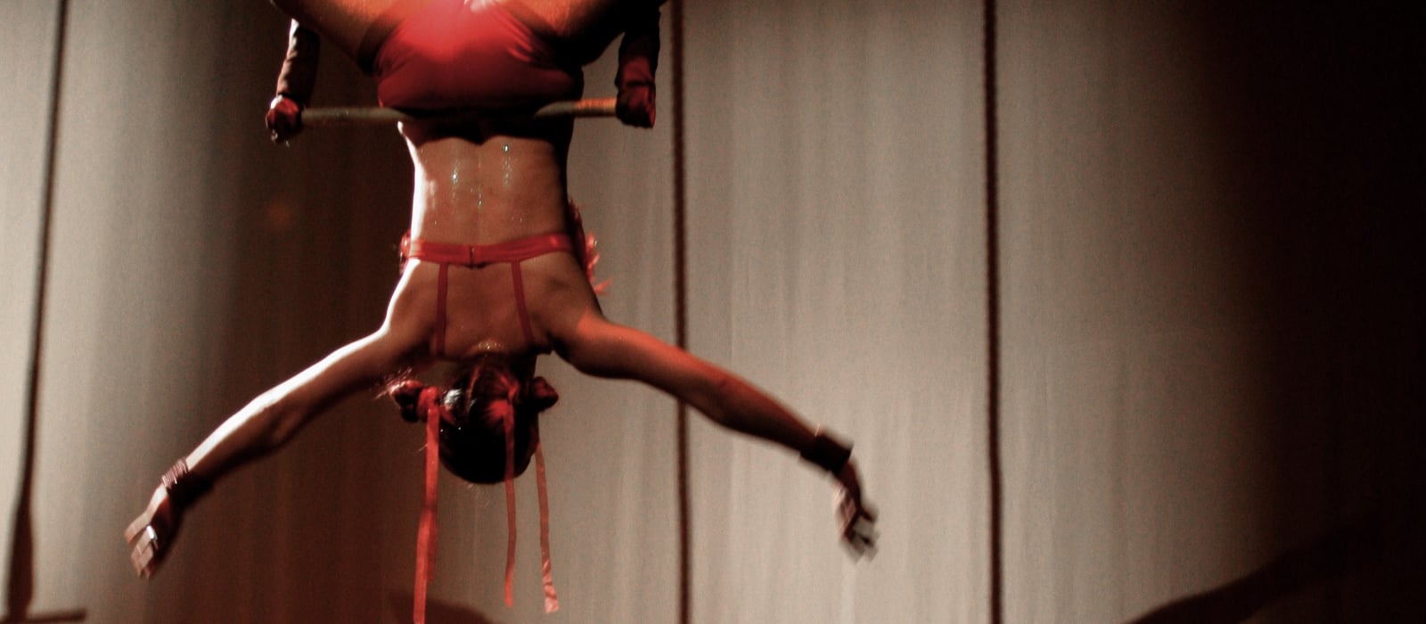 Aerialist hanging inverted from a trapeze in a red costume, arms open against a lit backdrop