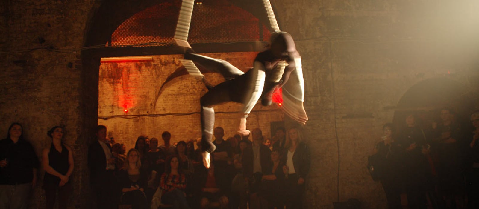Aerialist suspended in a white sling under the brick arches at Shunt, with the audience watching from the vault floor