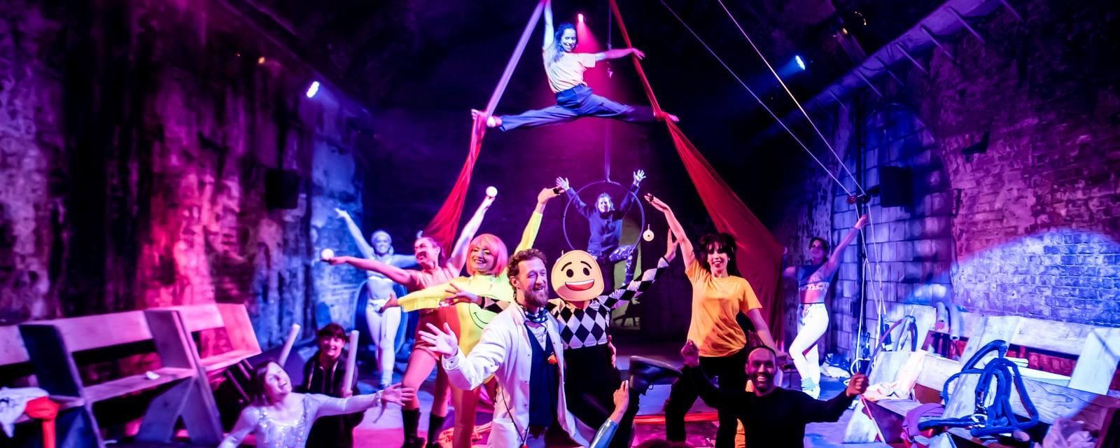 Happy Factory cast fill a brick vault: Director of Happiness, Moji in a yellow smiley mask, aerialists on silks and hoop under magenta light