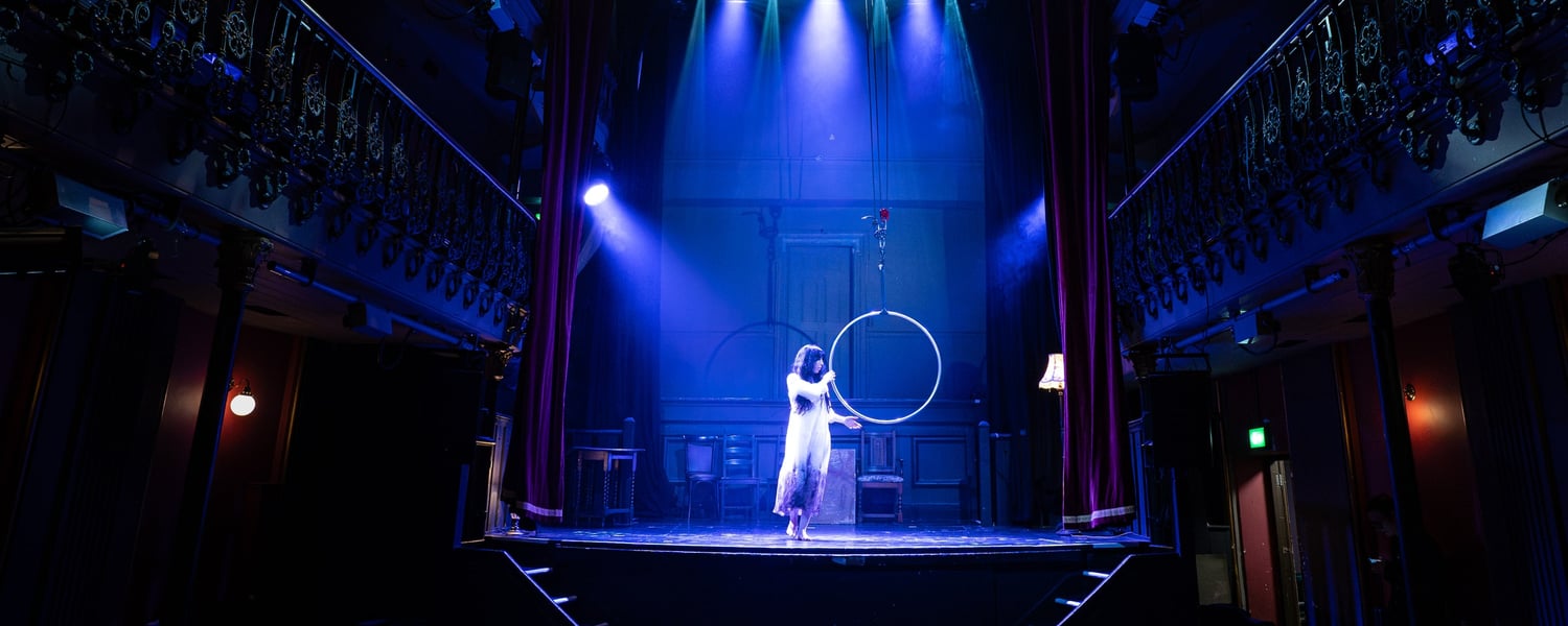 Wide view of Hoxton Hall: a performer in white stands centre stage beside an aerial hoop under blue spotlights